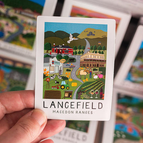 Hand holding a magnet with a colorful illustration of Lancefield, Macedon Ranges, featuring a train and buildings.