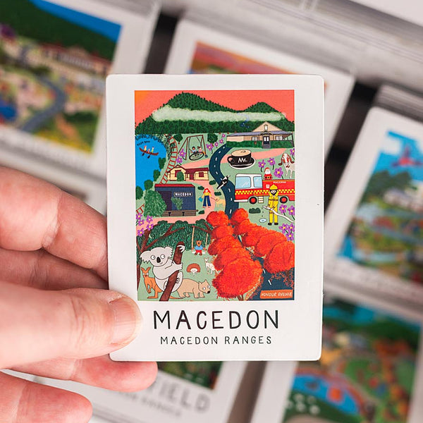 Colorful magnet with 'Macedon Ranges' text held by a hand, surrounded by other similar cards.