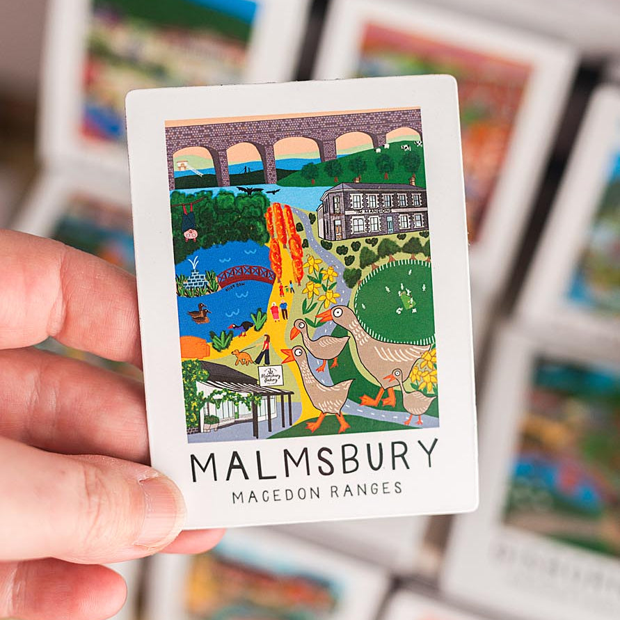 Hand holding a small magnet with a colorful illustration and 'Malmesbury' text, surrounded by more cards.