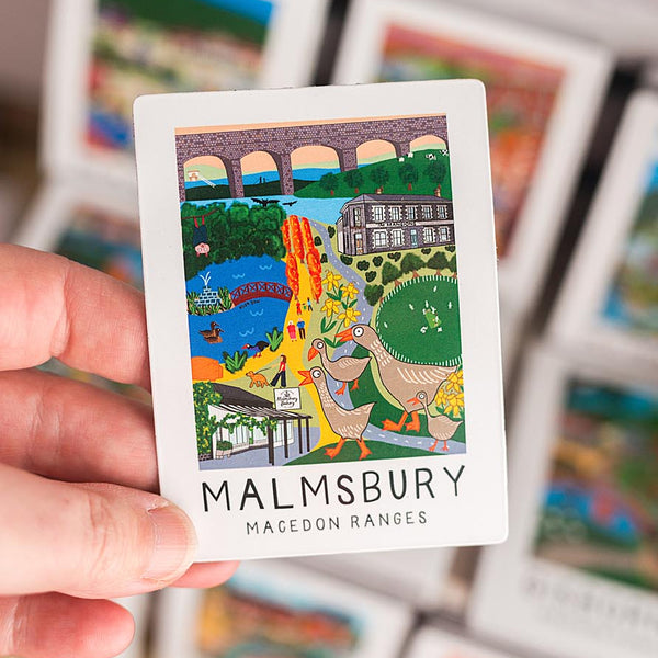 Hand holding a small magnet with a colorful illustration and 'Malmesbury' text, surrounded by more cards.