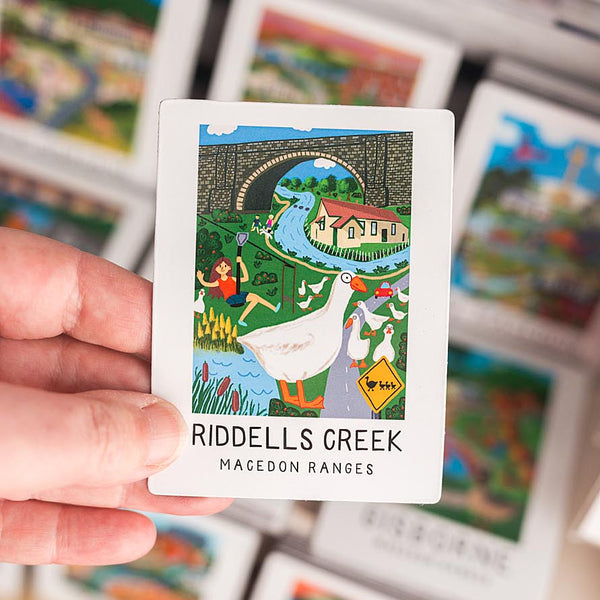 Hand holding a magnet with 'Riddells Creek' and 'Macedon Ranges' text, surrounded by other similar cards.