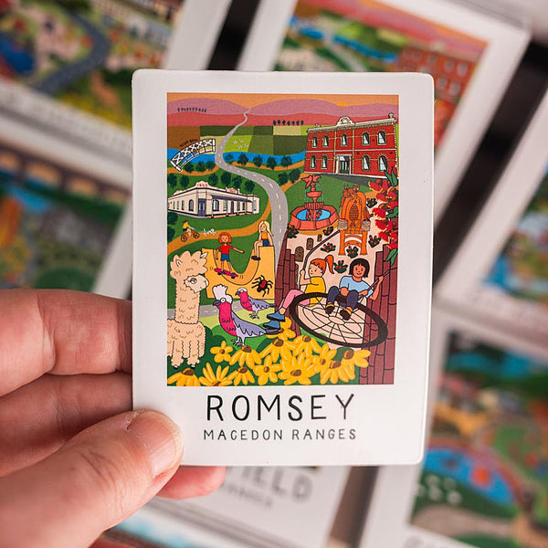 Hand holding a magnet with colorful illustration and 'ROMSEY' text, surrounded by other similar cards.