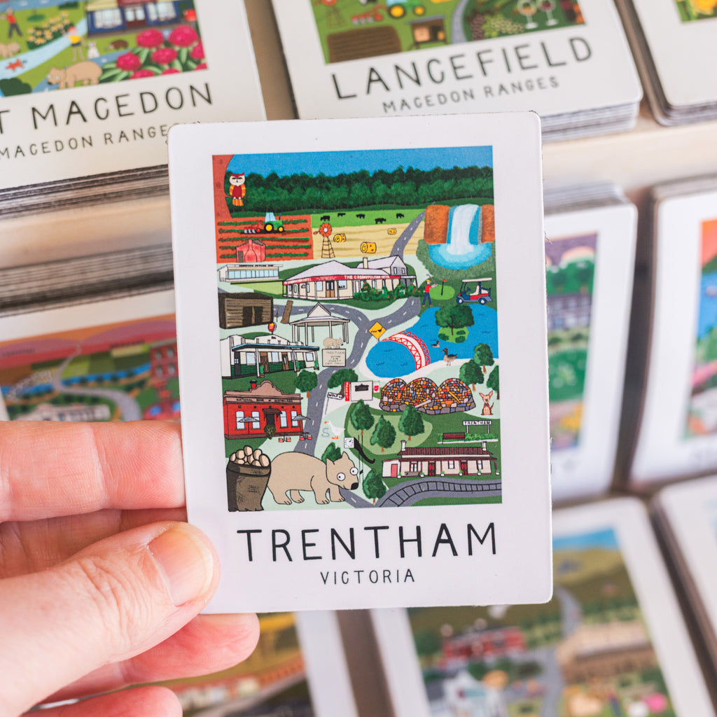 Card with a colorful illustration magnet of Trentham, Victoria held by a hand, with more cards in the background.
