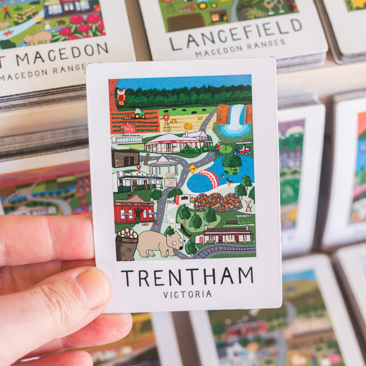 Card with a colorful illustration magnet of Trentham, Victoria held by a hand, with more cards in the background.