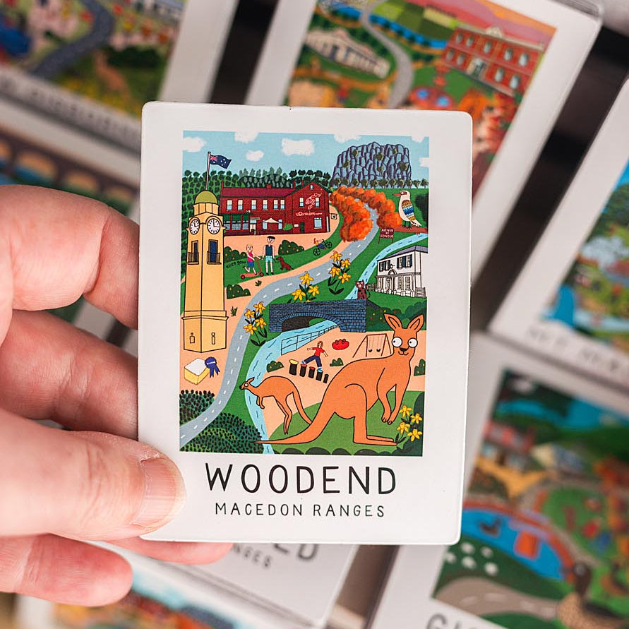 Hand holding a magnet with a scenic illustration and 'Woodend Macedon Ranges' text, surrounded by other similar cards.