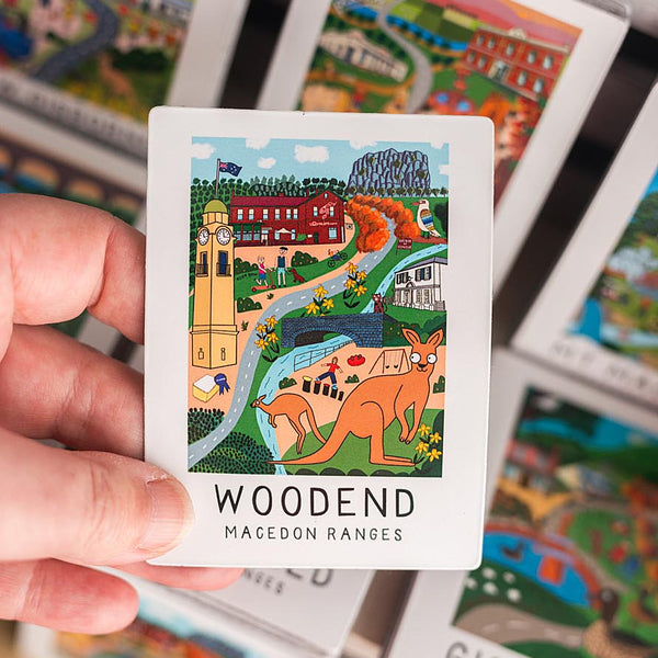Hand holding a magnet with a scenic illustration and 'Woodend Macedon Ranges' text, surrounded by other similar cards.