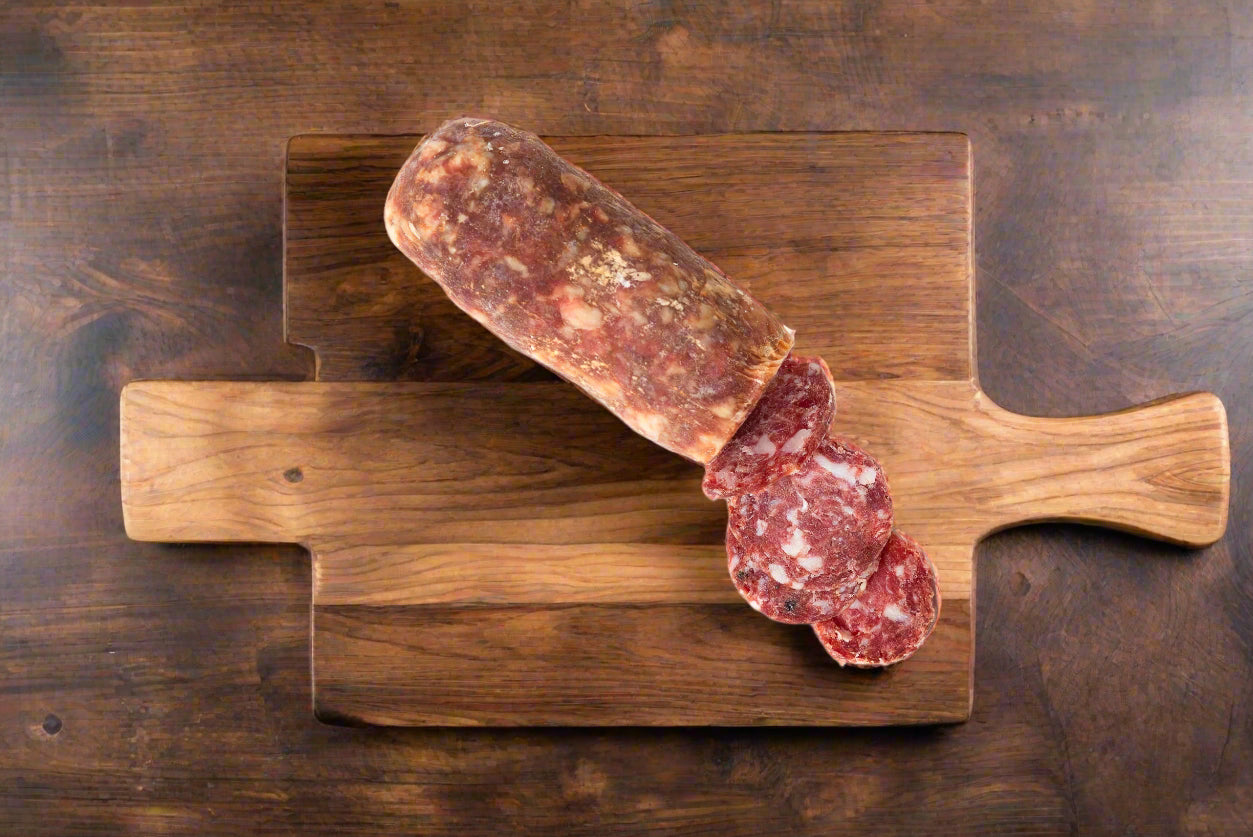 A whole stick of salami resting on a wooden cutting board, with a slice cut out to reveal the interior.